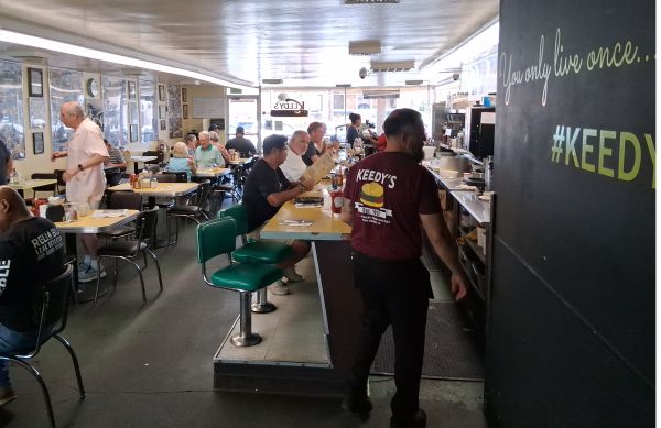 Interior of Keedy’s Fountain & Grill with green vinyl chairs, yellow tables, framed photos on the walls, and a server wearing a “Keedy’s Est. 1957” shirt.