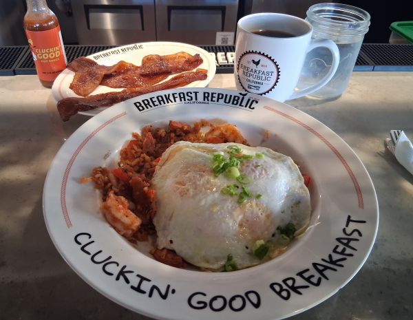 Breakfast Jambalaya topped with over‑easy eggs at Breakfast Republic in Palm Desert, served with Jurassic Pork bacon, coffee, and hot sauce.