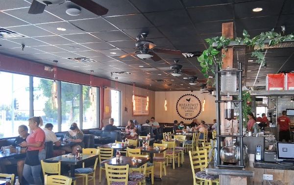 Wide interior view of Breakfast Republic in Palm Desert with yellow chairs, large windows, ceiling fans, and guests dining.