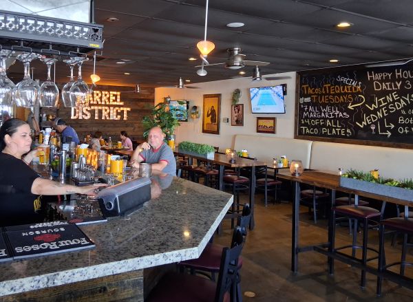 Interior of Barrel District featuring a granite bar, hanging glassware, patrons dining, warm lighting, and chalkboard specials.