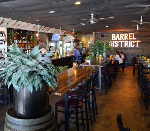 A modern rustic dining space with a long bar, wooden tables, potted plants, and patrons enjoying the restaurant.