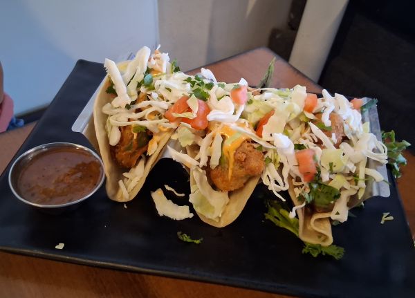 Three tacos filled with crispy avocado, shredded cabbage, tomatoes, cilantro, and sauce, served with a side of dipping sauce.