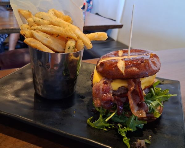 A pretzel‑bun cheeseburger with bacon, melted cheese, greens, and a side of seasoned fries served in a metal cup.