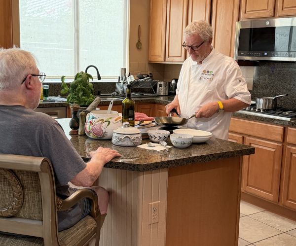 Chef Christophe cooking at a kitchen island while a senior client watches and engages in conversation