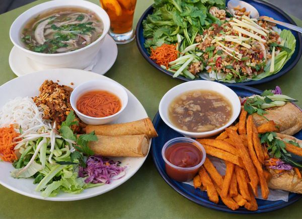 Four Vietnamese and Lao dishes — Pho, Vermicelli Bowl, Larb Salad, and Banh Mi — arranged on a green table for a shared meal.