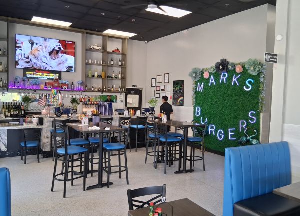 The modern interior of Mark’s Burgers featuring blue seating, a bar area, and the signature foliage wall with “Mark’s Burgers” lettering.