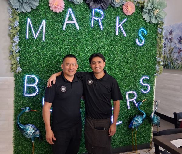 Mark and Marco Santana standing in front of the foliage wall and neon signage inside Mark’s Burgers in Cathedral City.