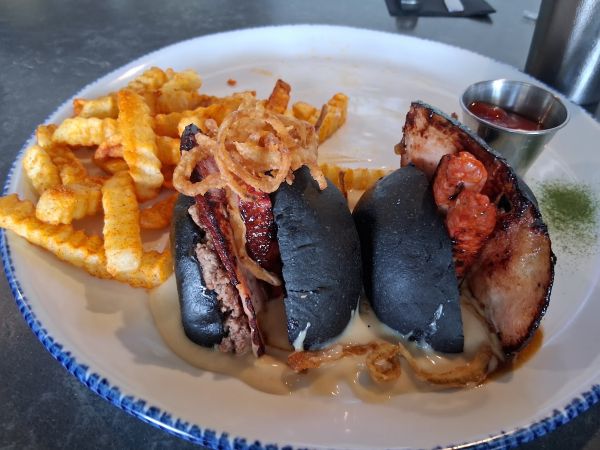 Mamo’s Super Hamburgesa served on a plate with a black brioche bun, crispy fried onions, and seasoned crinkle‑cut fries.