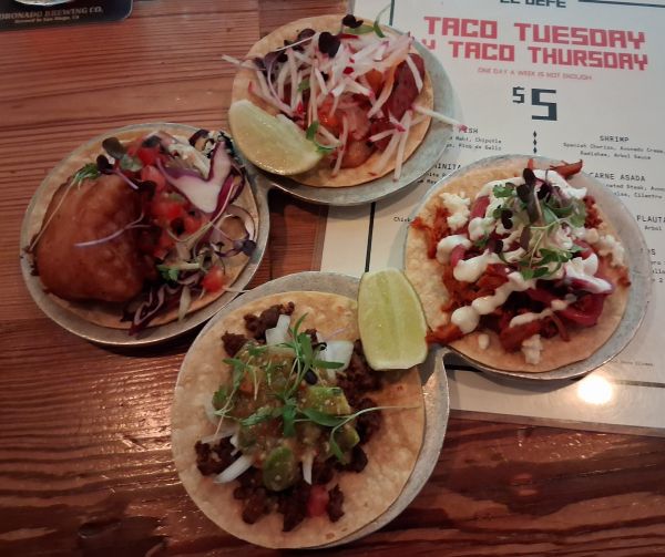Four gourmet tacos displayed on metal plates with the Taco Tuesday/Taco Thursday menu underneath.