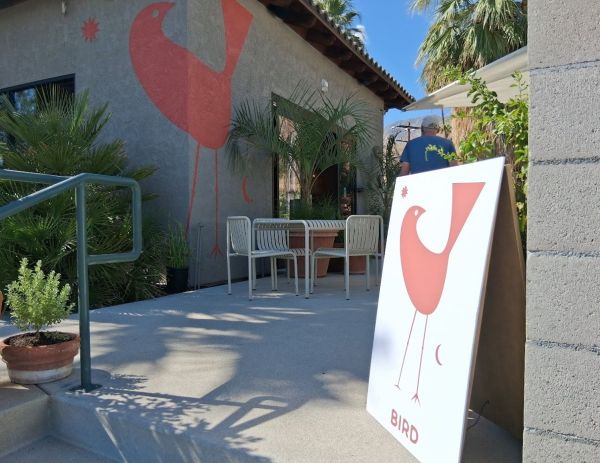 Exterior of Bird Palm Springs featuring the branded sandwich board sign and the large red bird mural on the building wall.