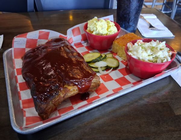 Tray of baby‑back ribs covered in barbecue sauce with coleslaw, potato salad, cornbread, pickles, and onions.