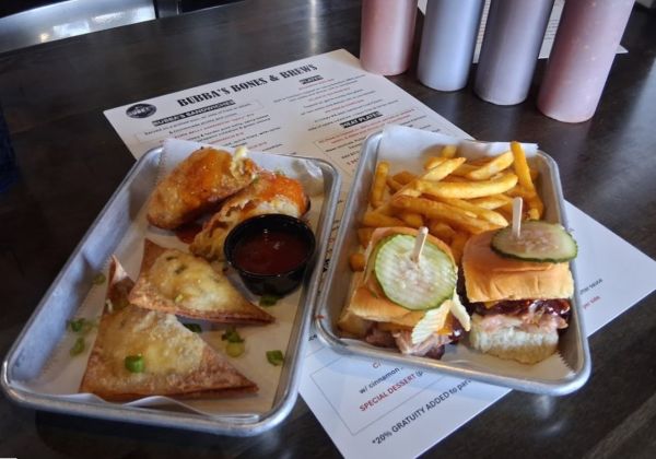 Trays of Bubba’s happy hour dishes including jalapeño sausage wontons, wings, brisket sliders with pickles, and fries, with house sauces in the background.