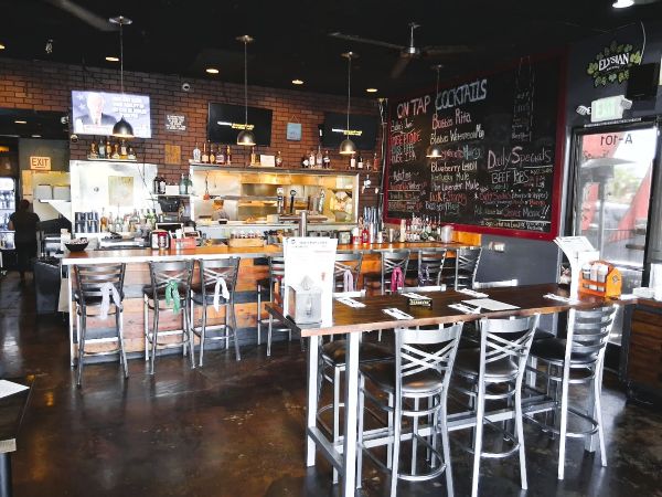 Interior of Bubba’s Bones and Brews showing the open kitchen, chalkboard drink menu, bar seating, and TVs above a brick wall.