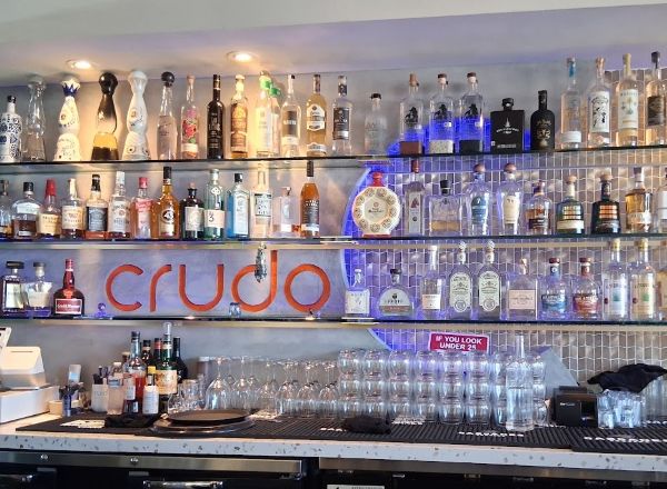 Shelves of spirits and glassware behind the bar with an orange Crudo sign centered on the wall