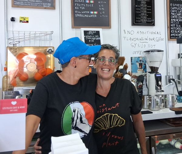 Candice and Paola smiling together behind the counter, wearing playful café shirts with Italian and French references.