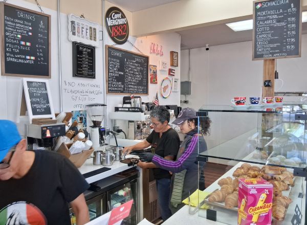 Candice and Paola standing behind the counter at Les Filles Café, surrounded by pastries, coffee equipment, and menu boards.