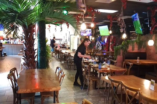 Long dining room at El Tranvia in Coachella with wood tables, palm trees, TVs and a server setting a table