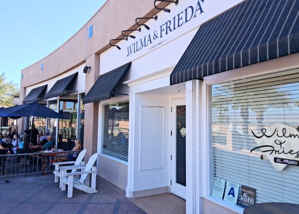 Wilma & Frieda storefront on El Paseo in Palm Desert with black awnings, patio tables and white Adirondack chairs.